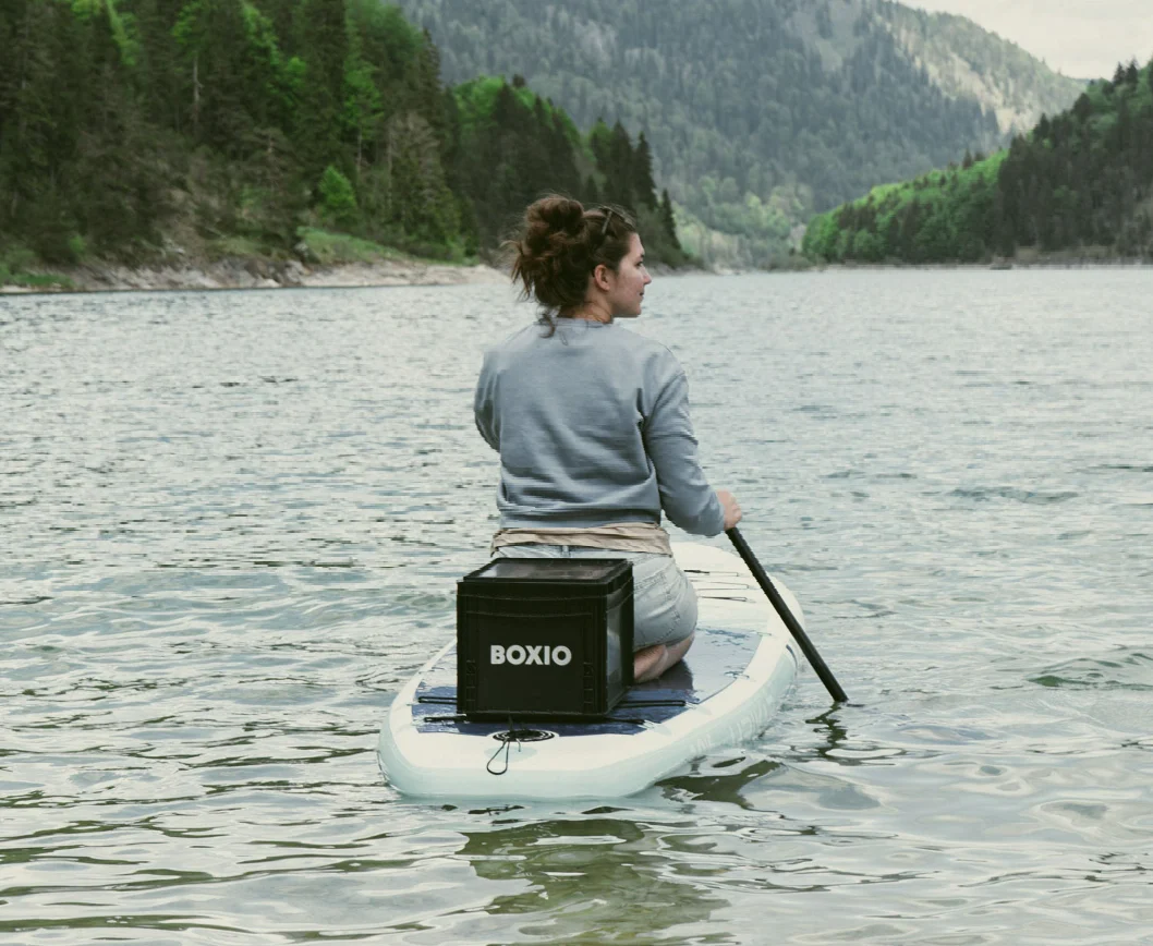 Frau paddelt auf einem Stand-Up-Paddleboard mit einem schwarzen BOXIO-Kasten in malerischer Berglandschaft.