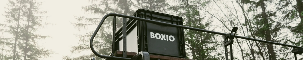 Schwarze Box mit dem Aufdruck "BOXIO" steht auf einem Gestell im Naturwald mit Bäumen im Hintergrund.