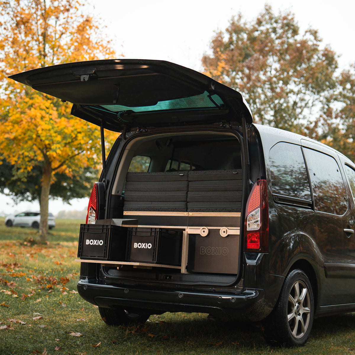 Heckansicht eines schwarzen Vans mit geöffnetem Kofferraum, ausgestattet mit Boxen und campingfreundlichen Möbeln.