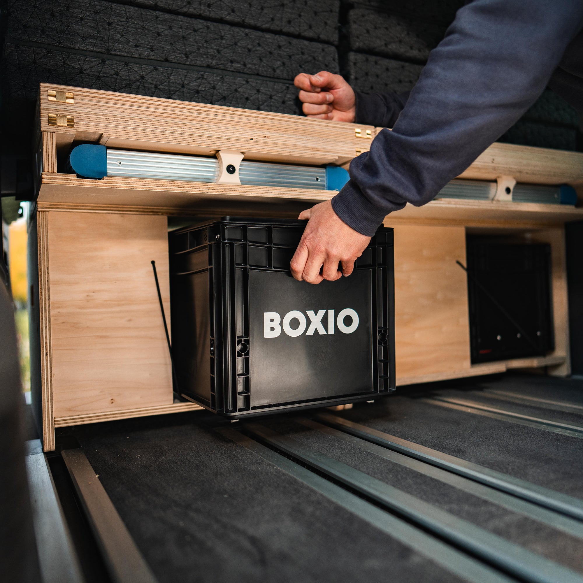 Person entnimmt eine schwarze BOXIO-Box aus einem Holzregal im Inneren eines Fahrzeugs.