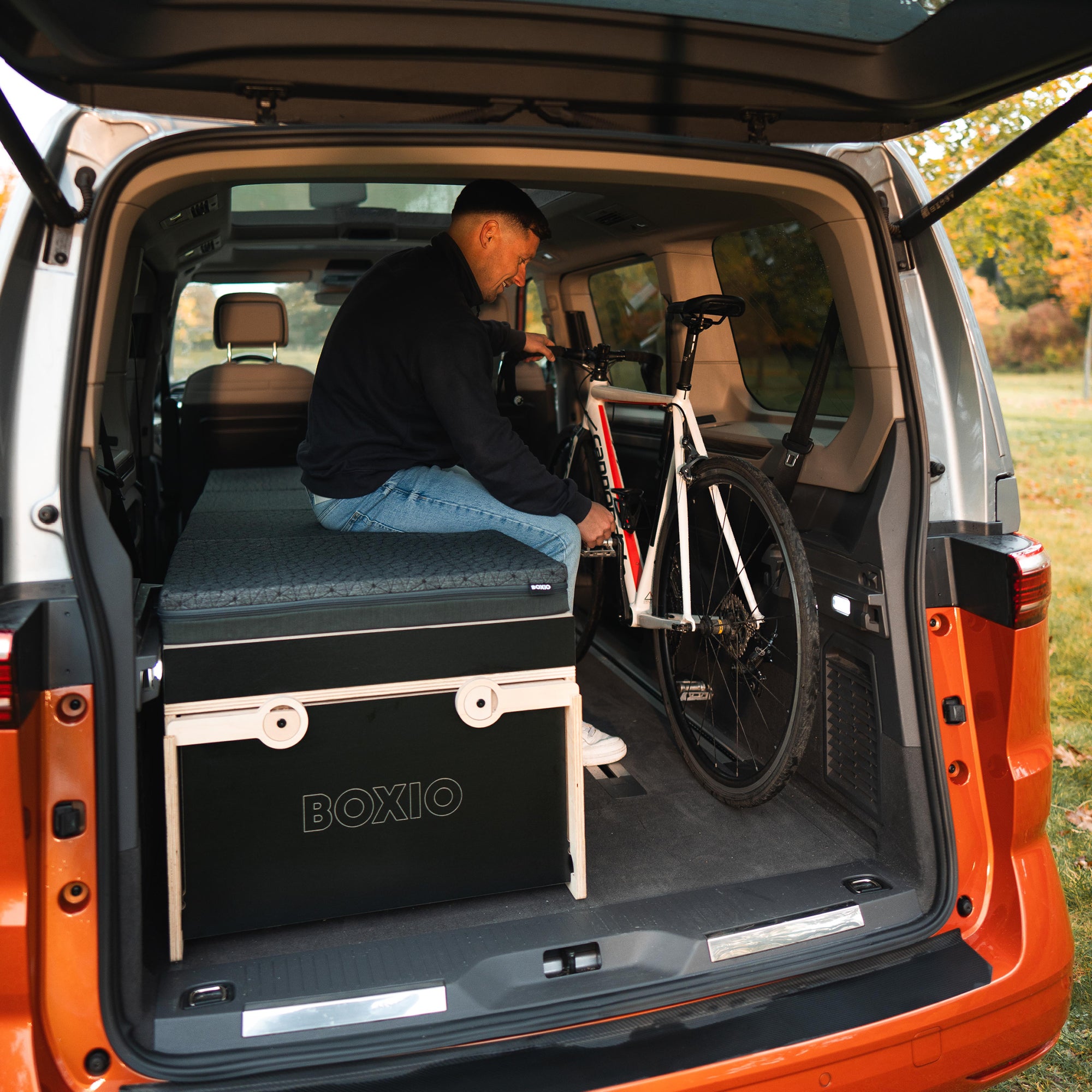 Mann sitzt im Kofferraum eines Autos und verstaut ein Fahrrad neben einer mobilen Box für Abenteuer.