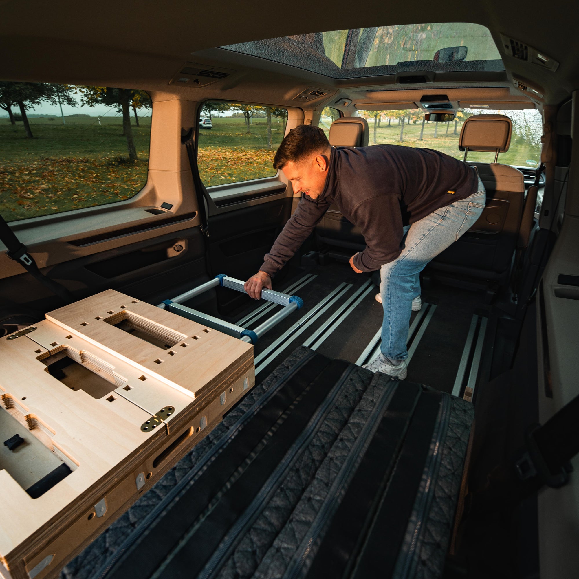 Mann montiert eine Holzstruktur im Innenraum eines Fahrzeugs mit modularer Sitzanordnung.