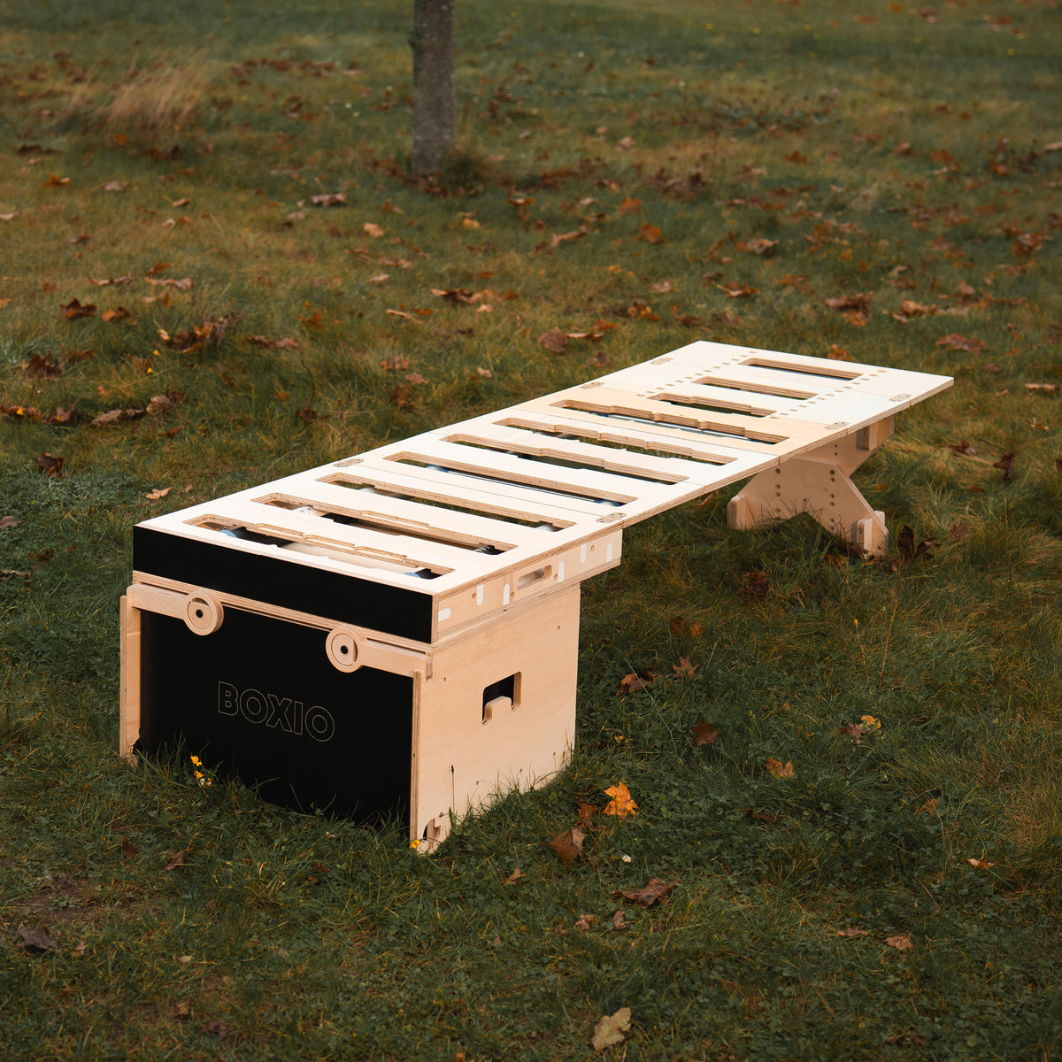 Holzbänke mit einem modernen, minimalistischen Design stehen auf einem grünen Rasen, umgeben von herbstlichen Blättern.