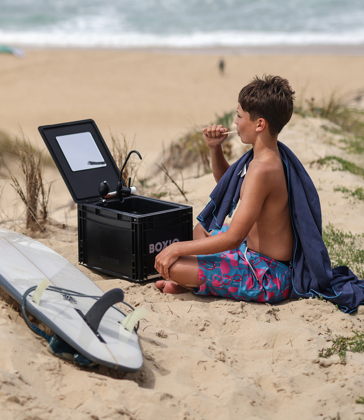 Junge im bunten Badeanzug sitzt im Sand, trinkt aus einer Box mit Spiegel, Surfbrett neben ihm, Strand im Hintergrund.