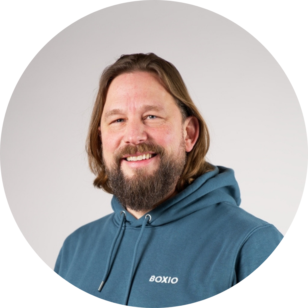Lächelnder Mann mit langem Bart in einem blauen Hoodie, präsentiert sich in runder Umrandung vor neutralem Hintergrund.